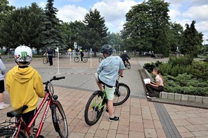 Na zdjęciu dwóch rowerzystów stojących przed torem przejazdu. W tle umundurowany policjant oraz dzieci jeżdżące na rowerach.