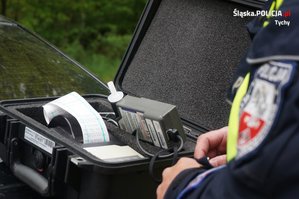 Na zdjęciu umundurowany policjant ruchu drogowego, który  stoi przy urządzeniu do badania stanu trzeźwości. Na urządzeniu widoczny wydruk.