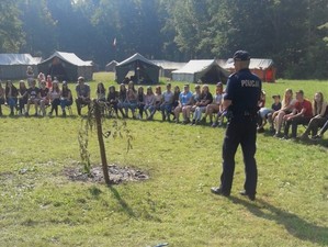 Na zdjęciu umundurowany policjant. Dookoła niego siedzą dzieci. W tle widać drewniane altanki.