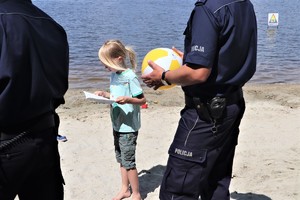 Na zdjęciu dwóch umundurowanych policjantów nad brzegiem jeziora. Pomiędzy nimi widać chłopca, trzymającego w rękach książeczkę. Jeden z policjantów trzyma dmuchaną piłkę plażową.