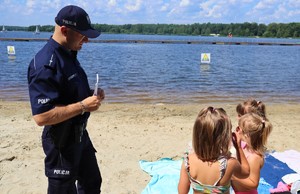 Na zdjęciu umundurowany policjant nad brzegiem jeziora. Przed policjantem trzy małe dziewczynki. Policjant trzyma w rękach książeczkę.