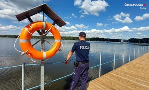 Na zdjęciu umundurowany policjant stojący na pomoście nad jeziorem. Obok policjanta koło ratunkowe.