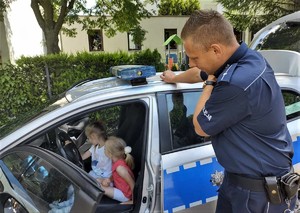 Na zdjęciu dwie dziewczynki siedzące za kierownicą policyjnego radiowozu. Obok radiowozu umundurowany policjant.