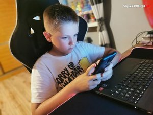 Na zdjęciu chłopiec siedzący przed komputerem z telefonem w rękach.