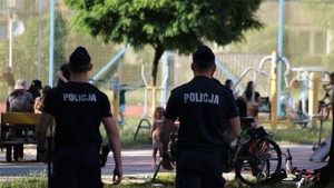 Na zdjęciu dwóch umundurowanych policjantów idących chodnikiem. W tle plac zabaw oraz dzieci.