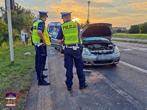 Na zdjęciu dwóch umundurowanych policjantów ruchu drogowego stojących na poboczu drogi. Przed nimi dwa uszkodzony samochód i karetka pogotowia ratunkowego.