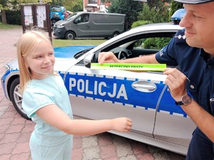 Na zdjęciu dziewczyna i policjant na tle radiowozu. Policjant trzyma odblask z napisem Bezpieczny pieszy.