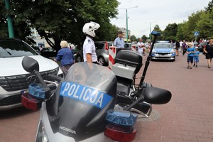 Na zdjęciu policyjny motocykl. W tle policjant z założonym kaskiem oraz radiowóz.