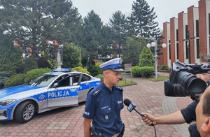 Na zdjęciu umundurowany policjant. Przed nim osoba trzymająca mikrofon i kamerę. W tle policyjny radiowóz.