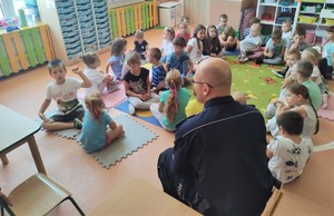 Na zdjęciu umundurowany policjant siedzący w sali pośród grupy dzieci w wieku przedszkolnym.