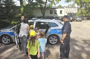 Na zdjęciu umundurowany policjant stojący obok radiowozu. Przy radiowozie grupa dzieci.
