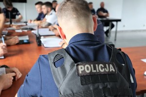 Na zdjęciu policjant w kamizelce z napisem Policja siedzący tyłem. W tle inni policjanci.