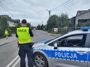 Na zdjęciu umundurowany policjant w kamizelce odblaskowej stojący obok radiowozu.