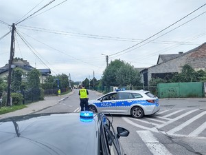 Na zdjęciu umundurowany policjant w kamizelce odblaskowej stojący obok radiowozu. Za nim radiowóz nieoznakowany z włączonym sygnałem świetlnym na dachu.