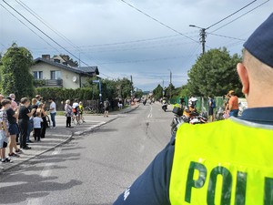 Na zdjęciu stojący tyłem policjant w kamizelce odblaskowej. Przed nim droga, po której jadą motocykle i pobocze, na którym stoją osoby.