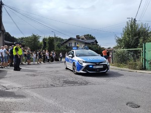 Na zdjęciu radiowóz pilotujący kolarzy. Obok skrzyżowania stoją policjanci.