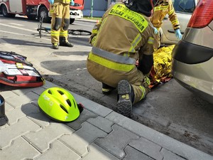 Na zdjęciu strażacy udzielający pomocy przedmedycznej. Obok nich na chodniku kask rowerowy.