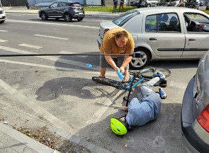 Na zdjęciu leżąca na jezdni rowerzystka. Obok niej mężczyzna zakładający rękawiczki.