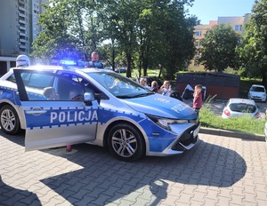 Na zdjęciu policyjny radiowóz. Obok niego dzieci oraz policjant.