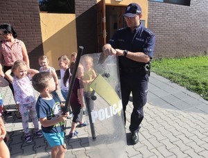 Na zdjęciu umundurowany policjant trzymający tarczę. Obok niego dzieci.