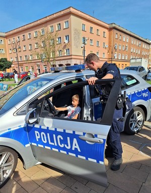 Na zdjęciu policyjny radiowóz oraz umundurowany policjant oraz chłopiec siedzący za kierownicą radiowozu.