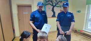 Na zdjęciu dwóch umundurowanych policjantów w szkolnej sali. Przed nimi dzieci wręczające podziękowanie.