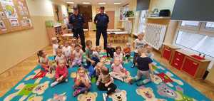Na zdjęciu dwóch umundurowanych policjantów w przedszkolnej sali. Przed nim siedzące na dywanie dzieci.