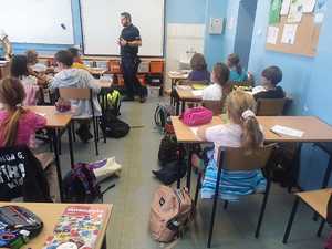 Na zdjęciu umundurowany policjant w szkolnej sali. Przed nim siedzący w ławkach uczniowie.
