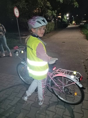Na zdjęciu dziewczynka stojąca przy rowerze, która ma założoną kamizelkę odblaskową z napisem Bądź widoczny