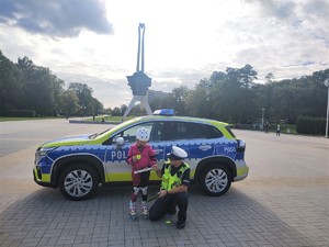 Na zdjęciu umundurowany policjant ruchu drogowego z dziewczynką na rolkach. Policjant wręcza dziewczynce odblask. Za nimi policyjny radiowóz.