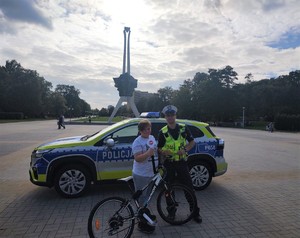 Na zdjęciu umundurowany policjant ruchu drogowego z chłopcem trzymającym rower. Za nimi policyjny radiowóz.