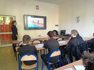 Na zdjęciu uczniowie klasy mundurowej w sali lekcyjnej. Przed nimi umundurowana policjantka oraz telewizor, na którym wyświetlany jest film.