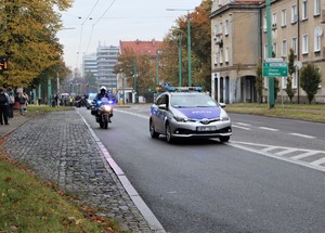 Na zdjęciu radiowozy i motocykl policyjny prowadzące przemarsz ulicami miasta.