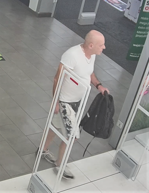 Na zdjęciu mężczyzna w wieku około 40 lat, wzrostu około 180 cm,  ubrany w białą koszulkę z krótkim rękawem, biało-czarne spodenki, ciemne buty, w ręce trzyma czarny plecak.