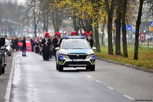 Na zdjęciu radiowóz policyjny. Za nim kolumna uczestników biorących udział w obchodach Dnia Niepodległości.