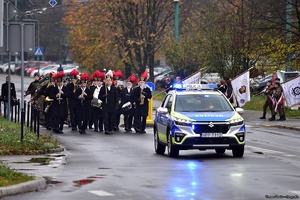 Na zdjęciu radiowóz policyjny. Za nim kolumna uczestników biorących udział w obchodach Dnia Niepodległości.
