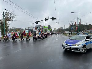 Na zdjęciu radiowóz policyjny. W tle widać kolumnę uczestników biorących udział w obchodach Dnia Niepodległości na czele z orkiestrą.