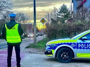 Na zdjęciu umundurowany policjant ruchu drogowego stojący przy radiowozie. W tle skrzyżowanie i jadące pojazdy.