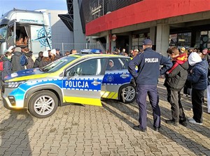 Na zdjęciu radiowóz policyjny i stojący obok niego umundurowany policjant. Dookoła osoby biorące udział w wydarzeniu.