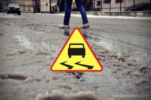 Na zdjęciu jezdnia z zalegającym śniegiem. Na środku ikona znaku drogowego oznaczającego: Uwaga slisko!