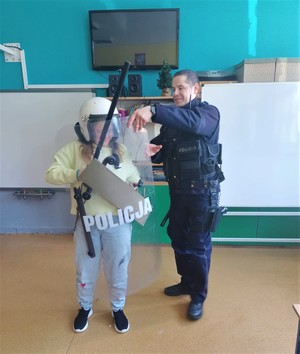 Na zdjęciu policjant oraz dziecko z założonym kaskiem, kamizelką, trzymające tarczę oraz pałkę.