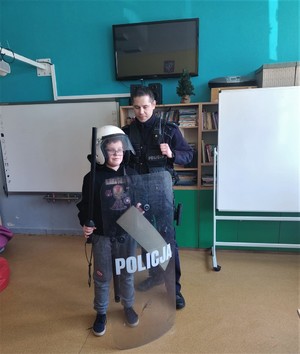 Na zdjęciu policjant oraz dziecko z założonym kaskiem, kamizelką, trzymające tarczę oraz pałkę.