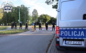 Na zdjęciu policyjny radiowóz. W tle umundurowani policjanci podczas zabezpieczenia meczu piłki nożnej.