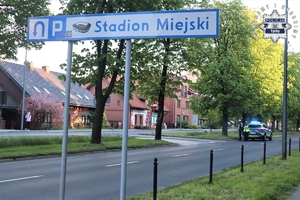 Na zdjęciu drogowskaz z napisem stadion miejski. W tle policjanci ruchu drogowego stojący obok radiowozu z włączonymi sygnałami świetlnymi.