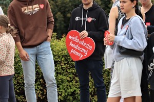 Na zdjęciu osoby, jedna z nich trzyma czerwone serce z napisem Wielka Orkiestra Świątecznej Pomocy.