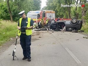 Na zdjęciu policjant ruchu drogowego. Za nim rozbity samochód i wozy straży pożarnej