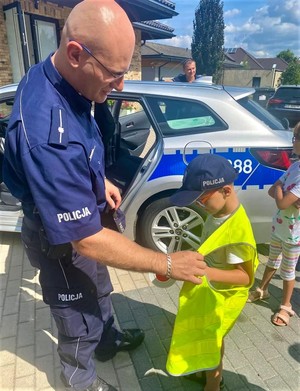 Na zdjęciu umundurowany policjant zakładający kamizelkę odblaskową chłopcu