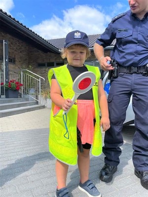 Na zdjęciu dziewczynka z założoną policyjną czapką oraz kamizelką odblaskową stojąca obok policjanta.