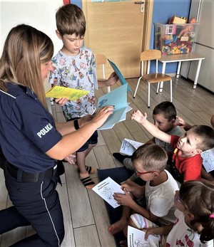 Na zdjęciu policjantka obok grupy dzieci. Policjantka trzyma w rękach laurki. Dzieci trzymają książeczki z napisem Elementarz Bezpieczeństwa.