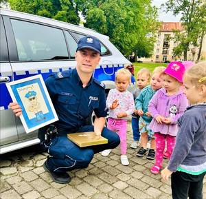 Na zdjęciu policjant z dziećmi trzymający ramkę z rysunkiem